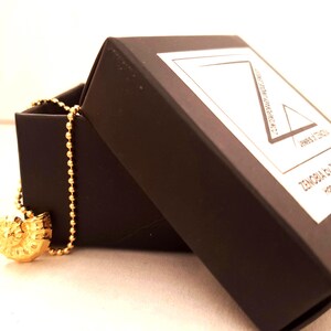 May include: A gold-colored pendant necklace with a spiral design, displayed in an open black jewelry box. The necklace has a ball chain. The box has a white label with the text "ZENOBIA by DENNEP ATES handmade jewelry".