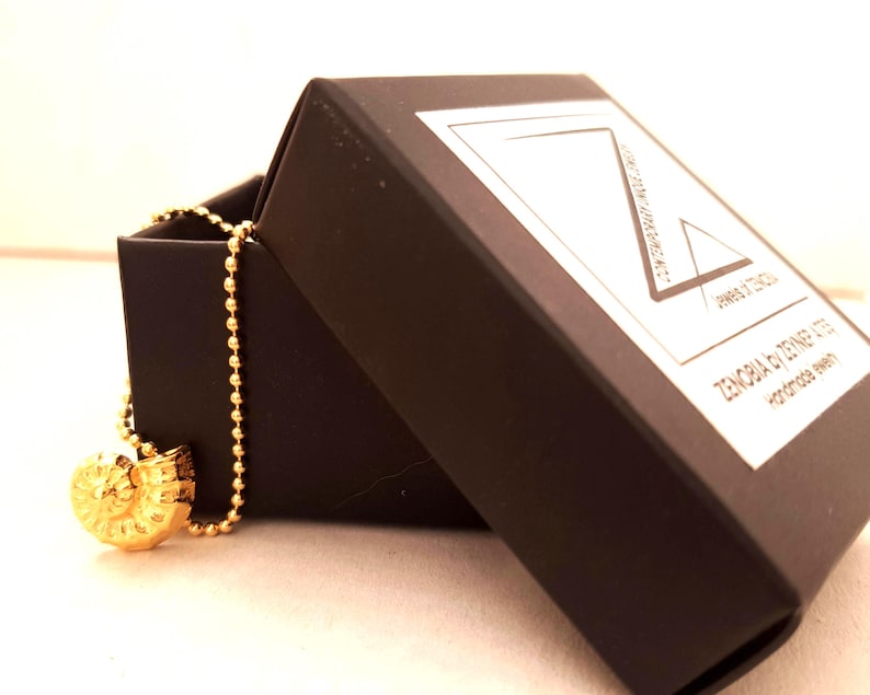 May include: A gold-colored pendant necklace with a spiral design, displayed in an open black jewelry box. The necklace has a ball chain. The box has a white label with the text "ZENOBIA by DENNEP ATES handmade jewelry".
