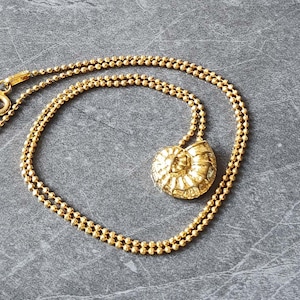 May include: A gold-colored necklace with a pendant shaped like a nautilus shell. The chain is made of small, round beads. The necklace is displayed on a gray surface.