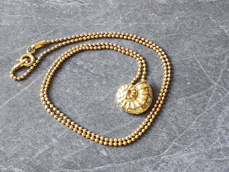May include: A gold-colored necklace with a pendant shaped like a nautilus shell. The chain is made of small, round beads. The necklace is displayed on a gray surface.