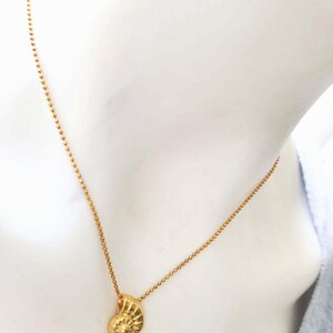 May include: A gold-colored necklace featuring a pendant shaped like a spiral seashell. The pendant hangs from a delicate chain of small, round beads. The necklace is displayed against a neutral background.