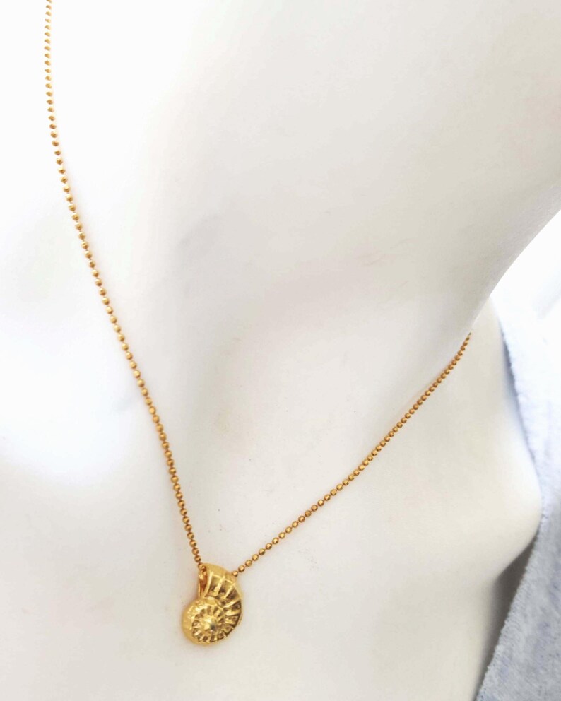 May include: A gold-colored necklace featuring a pendant shaped like a spiral seashell. The pendant hangs from a delicate chain of small, round beads. The necklace is displayed against a neutral background.