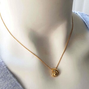 May include: A gold-colored necklace with a small, detailed pendant shaped like a nautilus shell. The necklace has a delicate chain of small, round beads. The pendant is approximately 0.5 inches in diameter. The necklace is displayed on a mannequin.