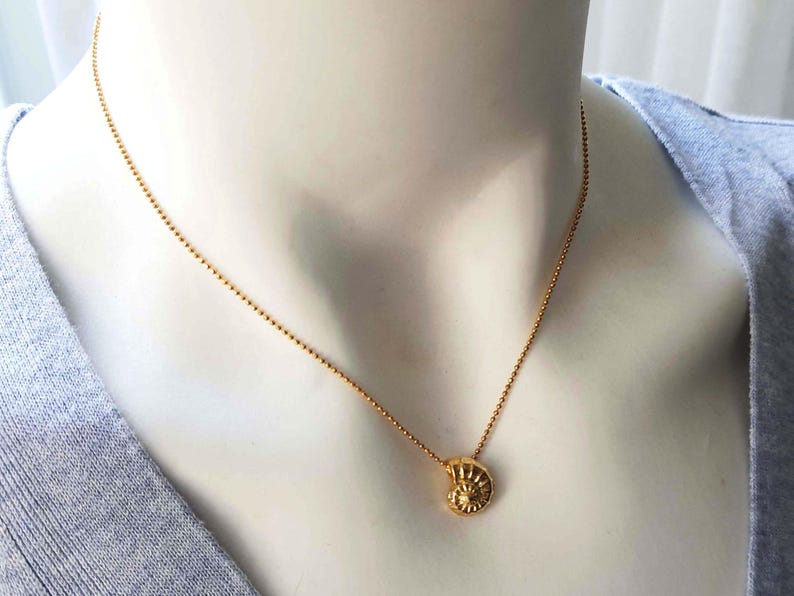May include: A gold-colored necklace with a small, detailed pendant shaped like a nautilus shell. The necklace has a delicate chain of small, round beads. The pendant is approximately 0.5 inches in diameter. The necklace is displayed on a mannequin.