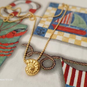 May include: A gold-colored necklace with a pendant shaped like a spiral seashell. The necklace is on a patterned background with nautical-themed illustrations, including a sailboat and a lobster.