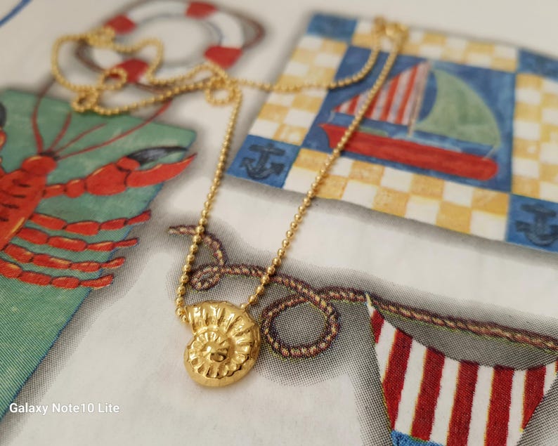 May include: A gold-colored necklace with a pendant shaped like a spiral seashell. The necklace is on a patterned background with nautical-themed illustrations, including a sailboat and a lobster.