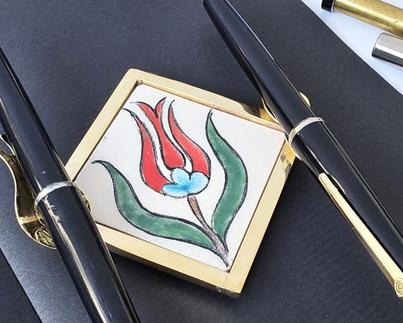 A handcrafted brass pen holder married to a hand-painted Turkish tulip ceramic rest — a writer's desk piece that feels like it belongs in an Istanbul bazaar, not an office supply aisle.