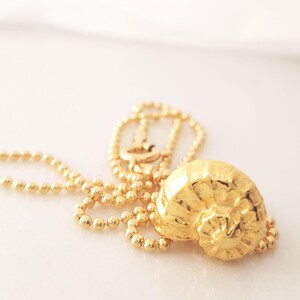 May include: A gold-colored necklace featuring a detailed, textured seashell pendant. The pendant is attached to a delicate ball chain. The necklace is displayed on a white surface, with soft lighting.