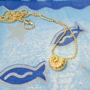 May include: A gold-colored necklace with a seashell pendant rests on a blue patterned surface. The necklace features a delicate chain and a detailed, spiral-shaped pendant. The background has a repeating pattern of fish and stars.