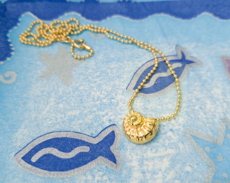May include: A gold-colored necklace with a seashell pendant rests on a blue patterned surface. The necklace features a delicate chain and a detailed, spiral-shaped pendant. The background has a repeating pattern of fish and stars.