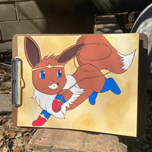 Può includere: Un portablocco con un personaggio dei cartoni animati di Eevee vestito da Wonder Woman. L'opera d'arte mostra un Eevee marrone con occhi blu, una fascia dorata e un costume rosso, bianco e blu. Lo sfondo è giallo chiaro.