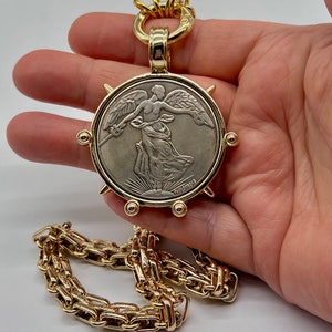 May include: A gold chain necklace with a large pendant featuring a silver coin with a winged figure and the inscription "PAOLOUBOIS".