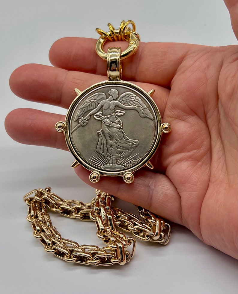 May include: A gold chain necklace with a large pendant featuring a silver coin with a winged figure and the inscription "PAOLOUBOIS".