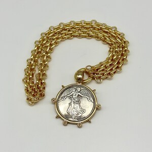 May include: A gold chain necklace with a large, round pendant featuring a silver-toned image of a winged figure. The pendant has a textured, circular border with six gold spikes.