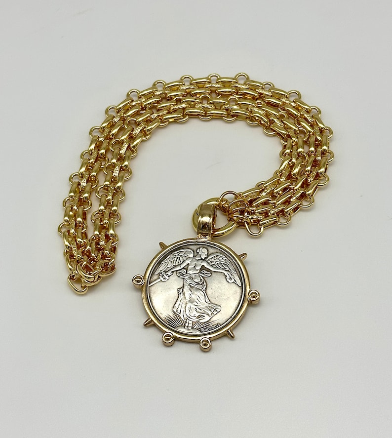 May include: A gold chain necklace with a large, round pendant featuring a silver-toned image of a winged figure. The pendant has a textured, circular border with six gold spikes.