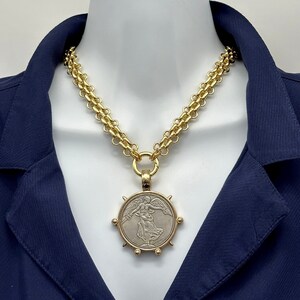 May include: A gold chain necklace with a large, round pendant featuring a silver-toned image of a winged figure. The pendant has a textured, circular border with gold accents.