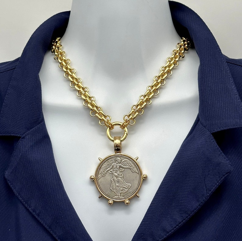 May include: A gold chain necklace with a large, round pendant featuring a silver-toned image of a winged figure. The pendant has a textured, circular border with gold accents.