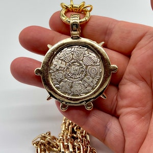 May include: A gold chain necklace with a large, round pendant featuring a circular design of multiple coats of arms. The pendant has a textured, spiky edge.