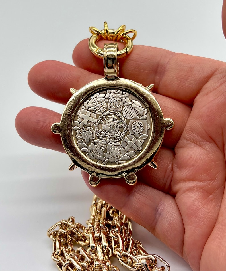 May include: A gold chain necklace with a large, round pendant featuring a circular design of multiple coats of arms. The pendant has a textured, spiky edge.