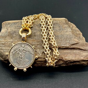 May include: A gold chain necklace with a pendant featuring a vintage silver coin with a winged figure. The coin is surrounded by a gold frame with spikes.