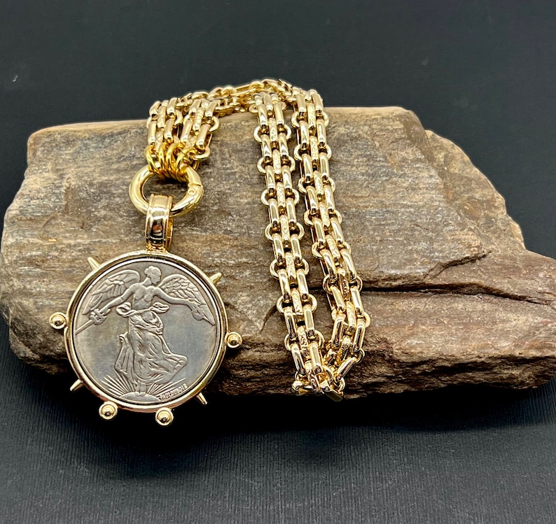 May include: A gold chain necklace with a pendant featuring a vintage silver coin with a winged figure. The coin is surrounded by a gold frame with spikes.