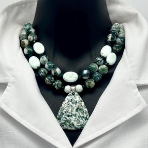 May include: A two-strand necklace with green and white beads and a triangular pendant. The pendant and some beads have a speckled pattern. The necklace is displayed on a black mannequin against a white shirt.