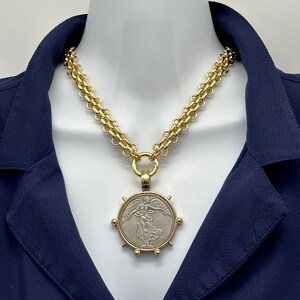 May include: A gold chain necklace with a large, round pendant featuring a silver-toned image of an angel with wings outstretched. The pendant is surrounded by a gold rim with small gold spikes.