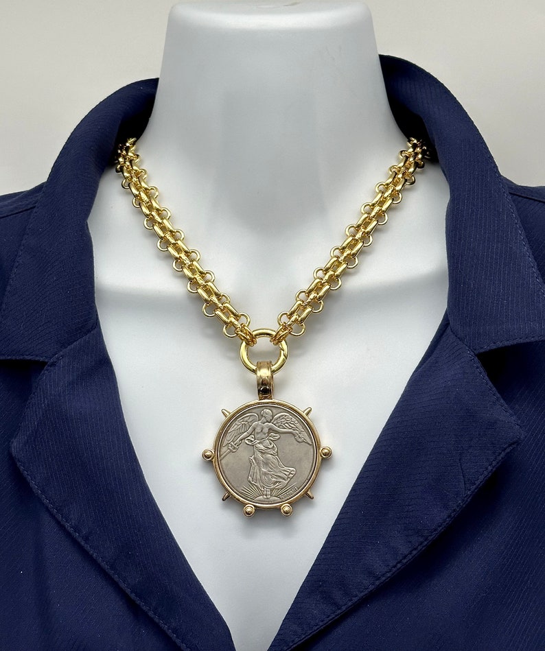 May include: A gold chain necklace with a large, round pendant featuring a silver-toned image of an angel with wings outstretched. The pendant is surrounded by a gold rim with small gold spikes.