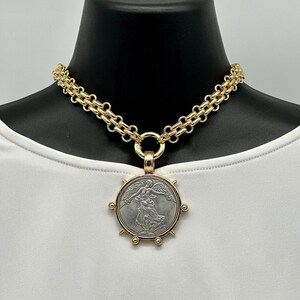 May include: A gold chain necklace with a large, round pendant featuring a silver coin with a winged figure. The pendant has a gold rim with spikes and small gold balls.