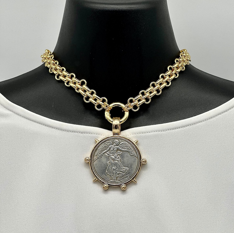May include: A gold chain necklace with a large, round pendant featuring a silver coin with a winged figure. The pendant has a gold rim with spikes and small gold balls.