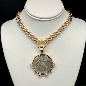 May include: A gold chain necklace with a large pendant featuring a silver coin with an image of a winged figure. The coin is surrounded by a gold frame with spikes.