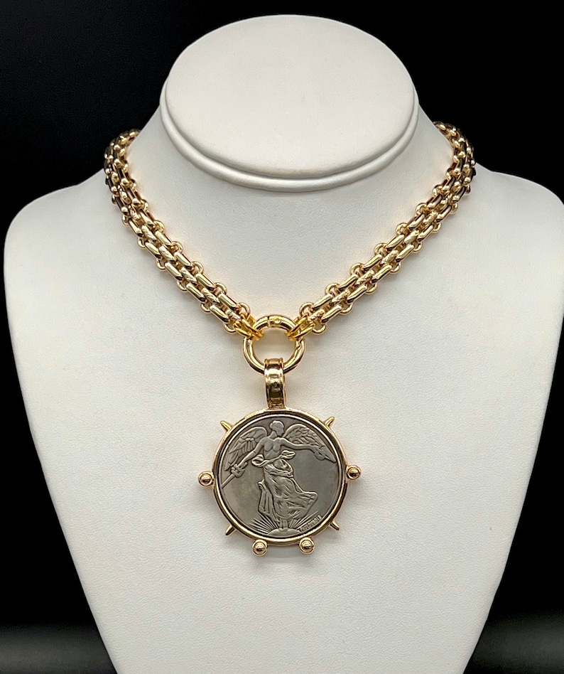 May include: A gold chain necklace with a large pendant featuring a silver coin with an image of a winged figure. The coin is surrounded by a gold frame with spikes.