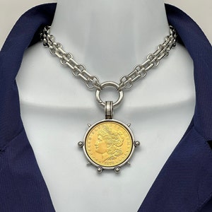 May include: A silver-toned chain necklace featuring a large, circular pendant. The pendant contains a gold-colored coin with the profile of a figure, the text E PLURIBUS UNUM, and the year 1878. The coin is set in a silver frame with small studs.