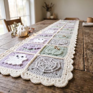Könnte beinhalten: Ein langer Häkel-Tischläufer mit einer Reihe quadratischer Motive in Pastelltönen wie Lavendel, Salbeigrün und Grau. Jedes Quadrat zeigt erhabene, strukturierte Designs, darunter weiße Geister, Spinnennetze und Kürbisse, mit einem gewellten Rand.