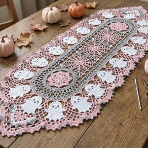May include: An oval crochet table runner featuring a central pink floral motif surrounded by a grey spiderweb pattern, bordered by white ghost figures and pink scalloped edges. A metal crochet hook and small scissors are placed nearby.