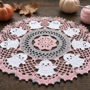 Op de afbeelding: Een rond gehaakt kleedje met een centraal roze bloemmotief omringd door een grijze cirkelvormige rand en een buitenste ring van acht witte spookfiguren met zwarte ogen en roze wangen, afgewerkt met een roze en grijze geschulpte kanten rand.