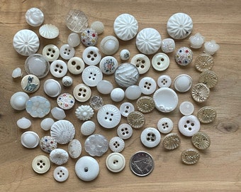 Op de afbeelding: Een verzameling witte en crèmekleurige knopen in verschillende vormen en maten, waaronder sommige met bloemmotieven. Een Canadese munt is ook in de afbeelding opgenomen.