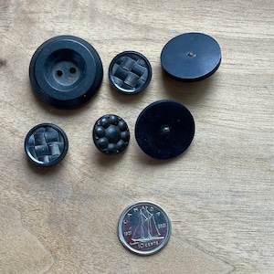 May include: A collection of seven black buttons, some with a textured design, and a Canadian 10-cent coin featuring a sailboat.