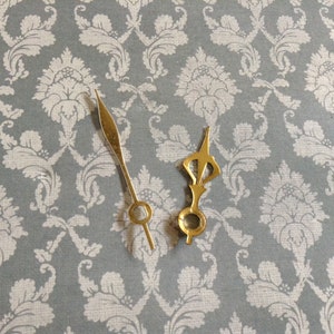 May include: Two gold clock hands, one with a simple pointed tip and the other with a decorative, ornate design. The hands are on a grey background with a white floral pattern.