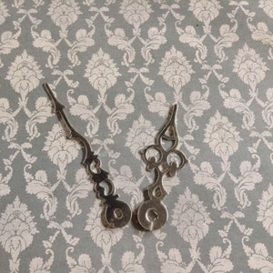 May include: Two silver clock hands with ornate designs. The hands are separated and appear to be for a clock face.