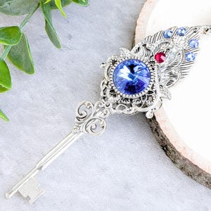 May include: Silver-toned key pendant necklace with a large, round, blue gemstone at the center. The pendant features intricate filigree and is adorned with smaller blue and red gemstones. The key design is detailed and elegant, perfect for a unique accessory.