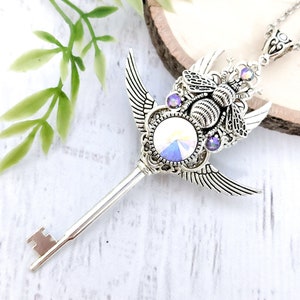 May include: A silver key necklace with a bee charm and iridescent crystal accents. The bee has wings and is surrounded by a decorative filigree design. The key has a long, slender shaft and a small, ornate handle.