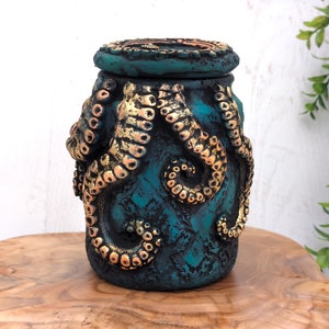 Könnte beinhalten: Eine tealblaue Keramikdose mit Deckel, verziert mit einem goldenen Kraken-Design. Die Dose hat eine strukturierte Oberfläche und einen Used-Look.