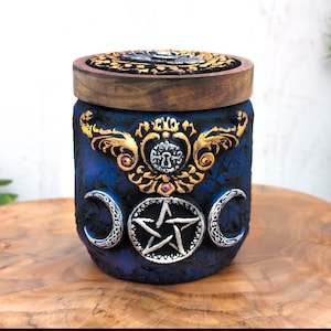 Peut inclure: Un pot décoratif bleu et or avec un couvercle en bois. Le pot présente un pentagramme, trois croissants de lune et un motif de trou de serrure.