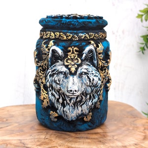 May include: A blue ceramic jar with a wolf's head design. The wolf's head is painted in white and silver with gold accents. The jar has a gold trim around the top.