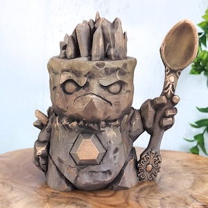 Könnte beinhalten: Eine kleine, braune, steinartige Figur mit einem kristallartigen Kopf und einem großen, goldenen Löffel in der Hand.