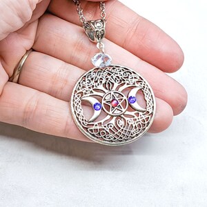 May include: Silver pendant necklace featuring a triple moon and pentacle design. The pendant showcases a silver Celtic knotwork design with three crescent moons and a pentagram at its centre. The pendant is embellished with red and blue gemstones.