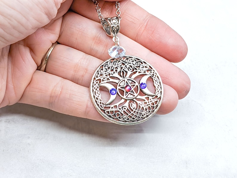 May include: Silver pendant necklace featuring a triple moon and pentacle design. The pendant showcases a silver Celtic knotwork design with three crescent moons and a pentagram at its centre. The pendant is embellished with red and blue gemstones.
