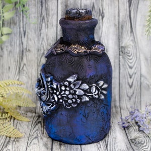 May include: A dark blue glass bottle with a cork stopper and intricate silver and gold floral designs. The bottle has a textured surface and is decorated with a swirling pattern.