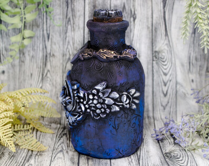 May include: A dark blue glass bottle with a cork stopper and intricate silver and gold floral designs. The bottle has a textured surface and is decorated with a swirling pattern.